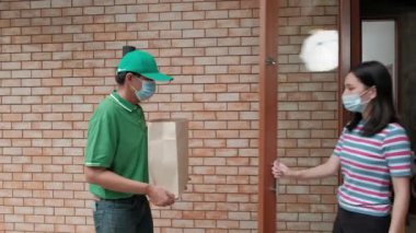 Yüzü maskeli yeşil üniformalı teslimatçı Asyalı kadının evine paket paketi veriyor. Çevrimiçi alışveriş, mobil uygulama üzerinden dijital ödemeler COVID-19 karantinasından yeni normal yaşam tarzı.