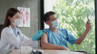 Asyalı erkek hasta, bir hastane kliniğinde koronavirüsü (COVID-19) aşıladığı sırada cep telefonuyla selfie portresi çekiyor. Enjeksiyonlar ateşi ve hastalığı tedavi eder..