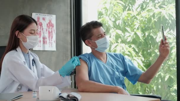 医師が予防接種をしている間に携帯電話で自撮り写真を撮るアジアの男性患者は 病院のクリニックでコロナウイルス Covid を予防します 注射は熱や病気を薬で治療します — ストック動画