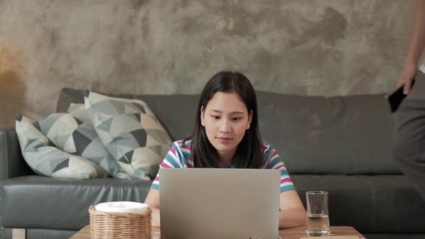 亚洲女人使用笔记本电脑进行网上交易 电子商务聊天和网络学习 她的哥哥坐在沙发后面 两个兄弟姐妹都在家里工作 因为他们必须与Covid 19隔离 — 图库视频影像