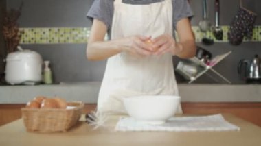 Bulanık ve yavaş çekimde, beyaz önlüklü kadın bir aşçı evin mutfağındaki ahşap bir masada yemek hazırlamak için bir yumurtayı fincana kırıyor. Yumurta sarısı yemek sağlıklı bir kahvaltıdır..