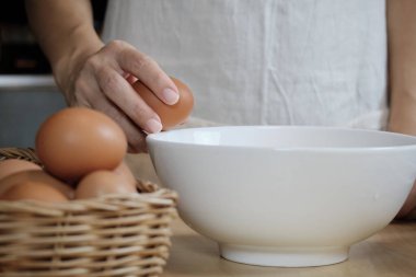 Yakın çekim görüntülerinde, beyaz önlüklü bir kadın aşçı ev mutfağındaki ahşap bir masada yemek hazırlamak için bir fincana yumurta kırıyor. Yumurta sarısı yemek sağlıklı bir kahvaltıdır..
