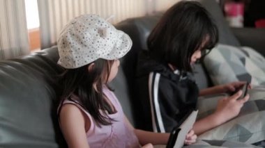 Gri koltukta oturan iki Asyalı (Taylandlı) kız, evin oturma odasında cep telefonlarıyla oyun oynuyorlar. Şapka takan abla dokunmatik ekranı kontrol etmek için tablet kullanıyor..