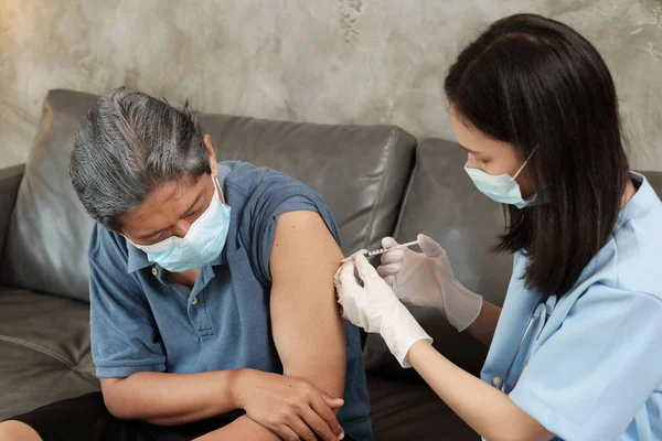 An Asian female doctor is vaccinating an elderly man who wears a mask to prevent illness from the virus infection. It is home healthcare by professionals to treat sick people who are retired.