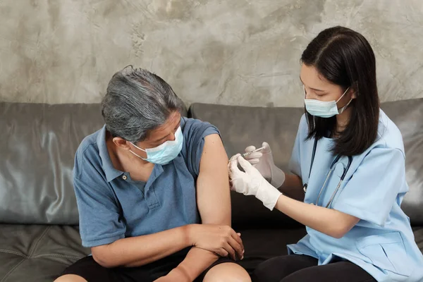 An Asian female doctor is vaccinating an elderly man who wears a mask to prevent illness from the virus infection. It is home healthcare by professionals to treat sick people who are retired.