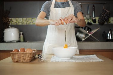 Yakın çekim görüntülerinde, beyaz önlüklü bir kadın aşçı ev mutfağındaki ahşap bir masada yemek hazırlamak için bir fincana yumurta kırıyor. Yumurta sarısı yemek sağlıklı bir kahvaltıdır..