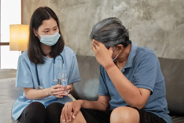 Female doctor with face mask holding a glass of drinking water while helping a male senior patient takes medicine. It is home healthcare, medical service for retired elderly people.