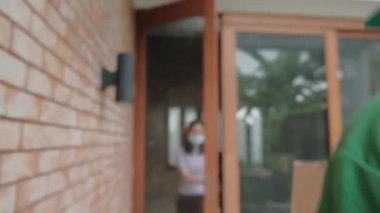 Yüzü maskeli yeşil üniformalı teslimatçı Asyalı kadının evine paket paketi veriyor. Çevrimiçi alışveriş, mobil uygulama üzerinden dijital ödemeler COVID-19 karantinasından yeni normal yaşam tarzı.