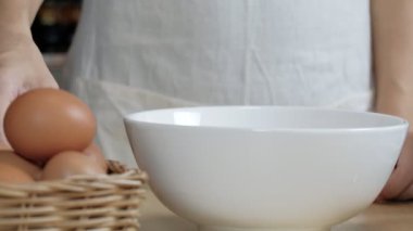 Yakın çekim görüntülerinde, beyaz önlüklü bir kadın aşçı ev mutfağındaki ahşap bir masada yemek hazırlamak için bir fincana yumurta kırıyor. Yumurta sarısı yemek sağlıklı bir kahvaltıdır..