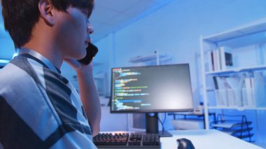 Genç Asyalı erkek bilişim programcısı ve teknoloji mühendisi uzaktan bilgisayar ve cep telefonu, uygulama geliştirme için yazılım kodları ve odadaki web ağı sistem verileri ile çalışıyor.