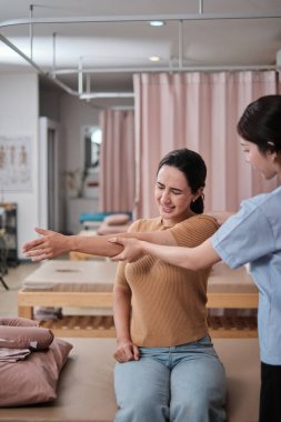 İspanyol kadın hasta kol esneme, tedavi, omuz kası ağrısı, Asyalı fizyoterapist tarafından tedavi, pasif tıbbi tedavi, sağlık masajı, spa kliniği.