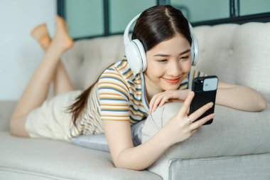 Güzel Asyalı kadın kanepede uzanıyor ve telefon kullanırken müzik dinliyor.