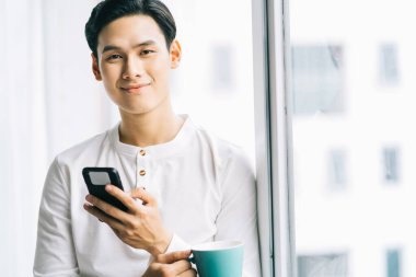 Asyalı işadamı mola sırasında kahve içiyor ve pencerenin kenarındaki telefona dokunuyordu.