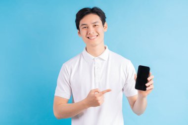 Gülümseyen genç adam elinde modern bir akıllı telefon tutuyor ve parmağını mavi arka planda izole edilmiş beyaz bir ekranda gösteriyor..