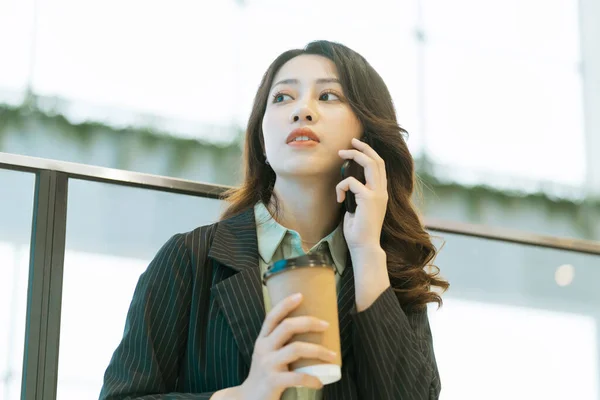 Portrait of a young Asian female director standing, drinking coffee and listening to the phone