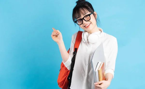 Asian female student wearing backpack behind her back, holding smartphone, neck wearing wireless earphones, holding paper cup