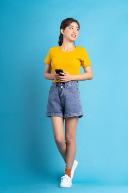 Genç Asyalı kadın ayakta duruyor ve mavi arka planda akıllı telefon kullanıyor.