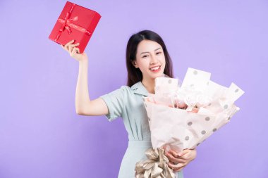 Genç Asyalı kadın elinde hediye kutusu ve mor arka planda çiçekler tutuyor.