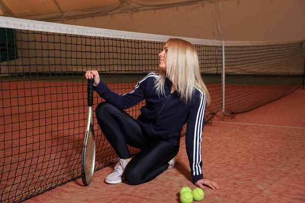 Young blonde woman with a tennis racket on an indoor tennis court with poor lighting