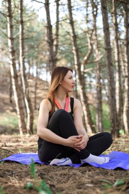 Genç bir kadın çam ormanında bir paspasın üzerinde egzersiz yapıyor.