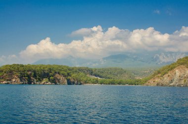 Güneşli bir günde yüksek dağlarla deniz kıyısı, dağların üzerinde bulutlar sallanıyor.
