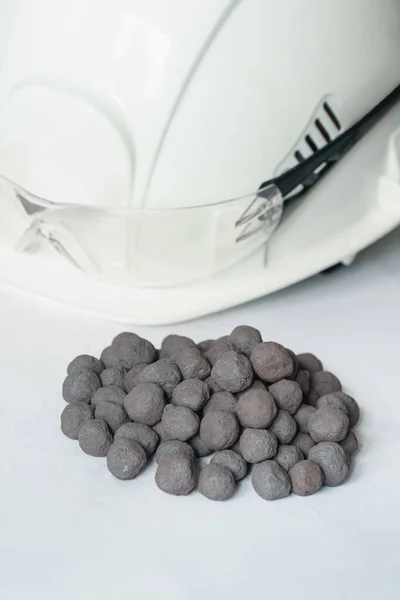 Iron ore pellets on a white background, with a white safety helmet and goggles in the background. Finished raw material for metallurgy iron ore granules.