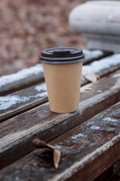 A cardboard disposable cup for tea or coffee stands on a bench board with melting snow in early winter or late fall.