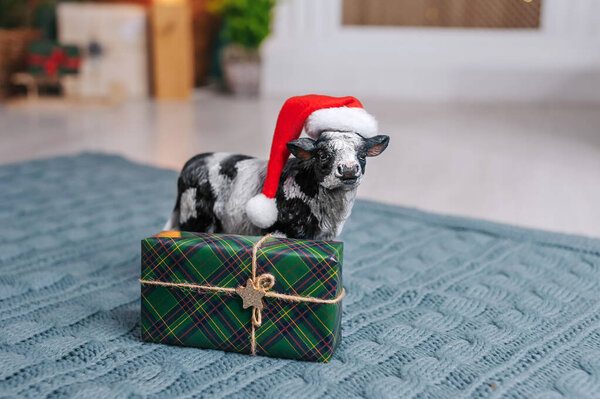 A cow in a New Year's hat is carrying a sleigh with gifts against the background of the interior
