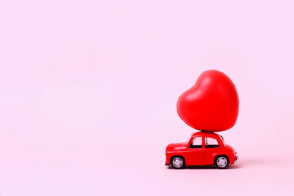 Маленький красный ретро-игрушечный автомобиль с сердцем на крыше на розовом фоне. Новый год, Рождество, День святого Валентина, Всемирный женский день. Продавцы скрывают пространство для копирования.