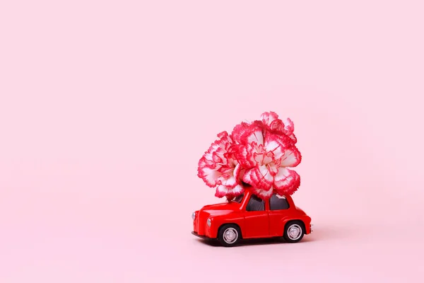 Small red retro toy car with a red and white flower on the roof against a pink background. Delivery of gifts for Valentine\'s Day, World Women\'s Day.