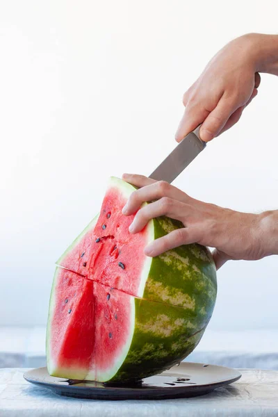 One hand holds ripe red watermelon, other holds knife and cuts off piece of watermelon. Vertical frame — Stock Photo, Image