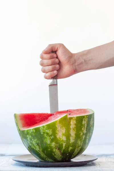 Half of red ripe watermelon on black plate on white background, hand sticks knife into watermelon. Vertical frame — Stock Photo, Image