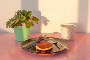 Pembe yüzeyde krepli siyah bir tabak var, yanında da bir çatal ve reçelli bir kaşık. Yanında bir fincan çay ve tencerede bir menekşe var. Kahvaltı konsepti ve günaydın