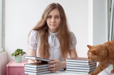 Mavi gözlü ve kahverengi saçlı bir kız masada oturuyor, önünde iki yığın kitap var, bir kitabı elleriyle tutuyor, yanında bir kedi var..