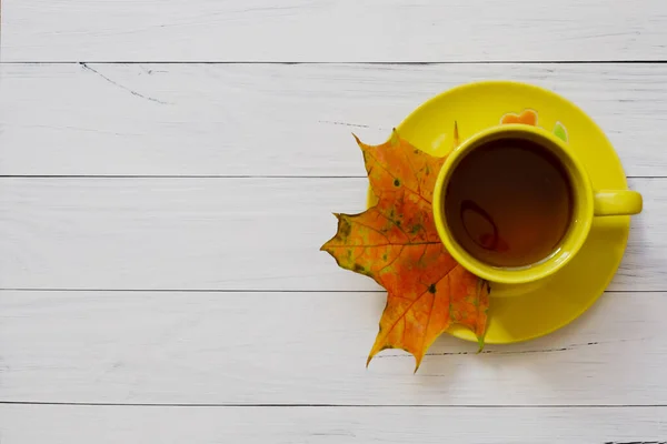 Yellow mug and autumn maple leaf on wooden background, flat lay
