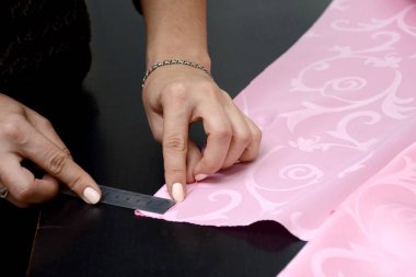 Kadının elleri, pembe kumaşı döndürmek için gerilmeyi ölçer. Gri metal cetvelli bir ölçü. Stüdyodaki siyah bir masaya perde dikme hazırlığı. Küçük işletme kavramı.