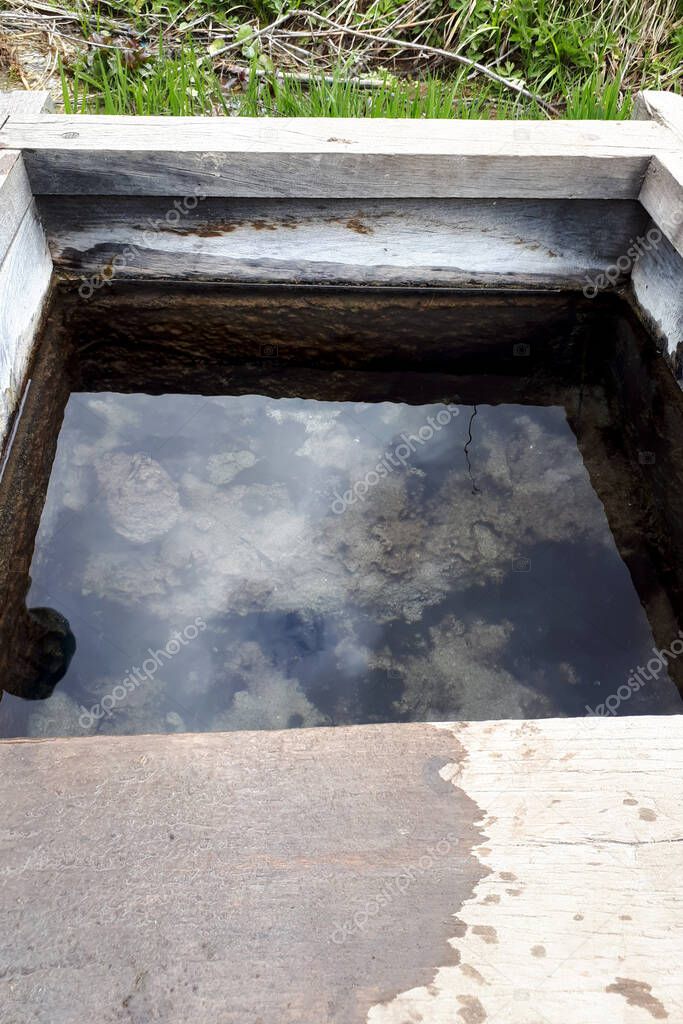 Wooden well with clear water in the countryside. Reflection of the sky with clouds in calm water. Vertical photo.