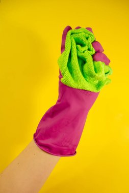 Sarı arka planda açık yeşil bir bez tutan pembe lastik eldivenli bir el. Ev temizliği ve temizlik konsepti
