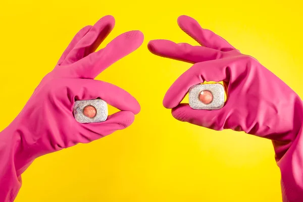 Pembe eldivenli kadın elleri sarı arka planda bulaşık deterjanı tabletleri tutuyor. Evdeki temizlik kavramı.