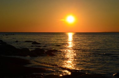 Gölün üzerinde parlak gün batımı. Güneş ormanın arkasında battı. Sarı günbatımı. Karanlık suda siyah taşlar. Kaya kıyısı.