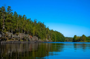 Mavi nehir ve açık mavi gökyüzü. Kıyılar boyunca yeşil çam ormanı ve sarı huş ağaçları vardır. Kayalık sol kıyısı suya yansıyor..