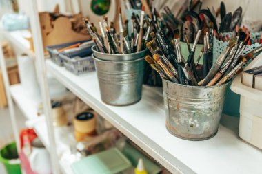 Metal kovalarla resim yapmak için birçok fırça bir sanat stüdyosundaki beyaz ahşap raflarda durur.