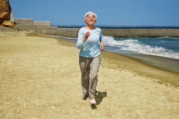 Пожилая счастливая женщина бегает по пляжу вдоль побережья у моря — стоковое фото