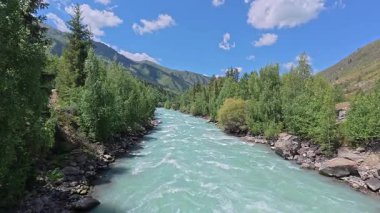 Koksu Nehri, Kazakistan 'daki manzaralı bir dağ vadisinde, parlak mavi gökyüzünün altında, yumuşak bulutlarla akıyor. Turkuaz su, kayalık kıyılar ve yoğun yeşil orman Orta Asya 'nın el değmemiş doğal bir manzarasını yaratır..
