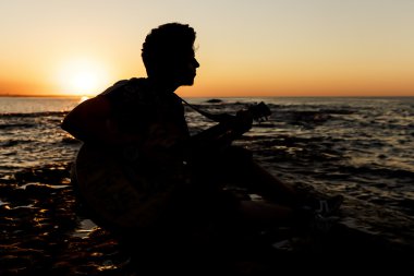 Genç adam (yumuşak odak günbatımında bir gitar çalmak)