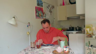 Bir adam sıcak bir mutfak masasında oturmuş kahvaltısının tadını çıkarırken çeşitli yiyecek ve içeceklerle etkileşime geçiyor ve sabah deneyimine biraz keyif katıyor.