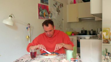 Sıcak bir mutfak masasında oturan bir adam ev dekorasyonları ve mutfak aletleriyle çevrili lezzetli makarna ve içeceklerin tadını çıkararak rahat bir yemek atmosferi yaratıyor.