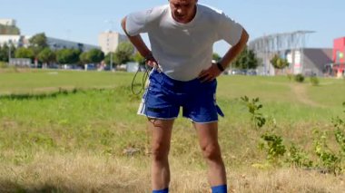 Spor kıyafeti giymiş bir adam parkta koştuktan sonra esniyor. Rahatsızlığı azaltmak ve esnekliğini arttırmak için egzersiz yapıyor. Formuna ve sağlığına katkıda bulunuyor.