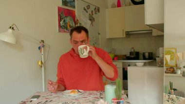 Sıcak bir mutfakta, rahat bir masada oturan bir adam kahvaltısını yaparken sıcak bir fincan kahvenin ve lezzetli bir aperatifin tadını çıkarıyor. Arka planda sayısız mutfak malzemesi görülebilir.