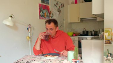 Beyaz adamın mutfak masasında çayını yudumlaması, renkli masa örtüsü, kupa ve tabak, sıradan kapüşonlu, eskiz kitabı ipuçları, yumuşak sabah ışığı, tefekkür ifadesi, ev sakinliği.